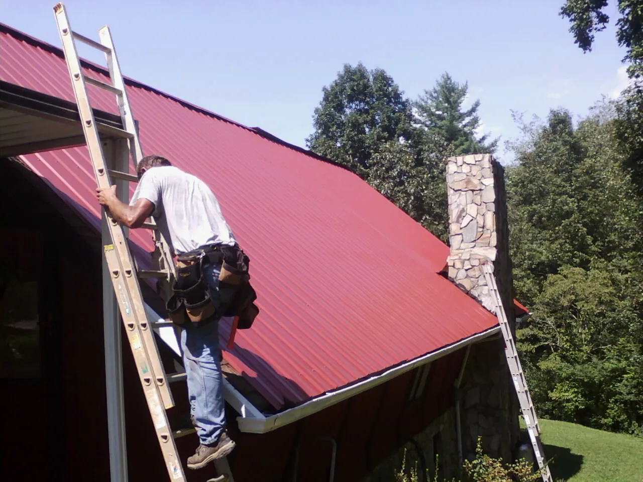 Roofer on a ladder beside a red metal roof and stone chimney — Soffit & Fascia Repair by Roofing Brothers in Nolanville